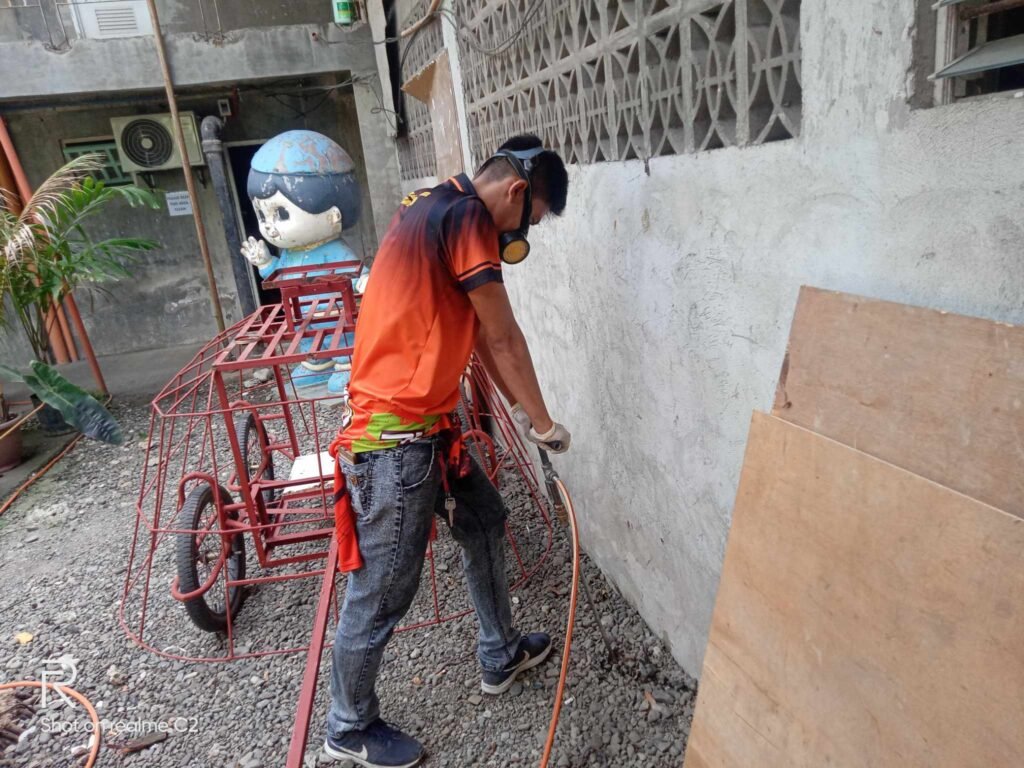 Technician from Jansen Pest Control Boracay performing pest control treatment near a concrete wall, wearing protective gear and handling equipment, with safety materials and a red containment cart in the background