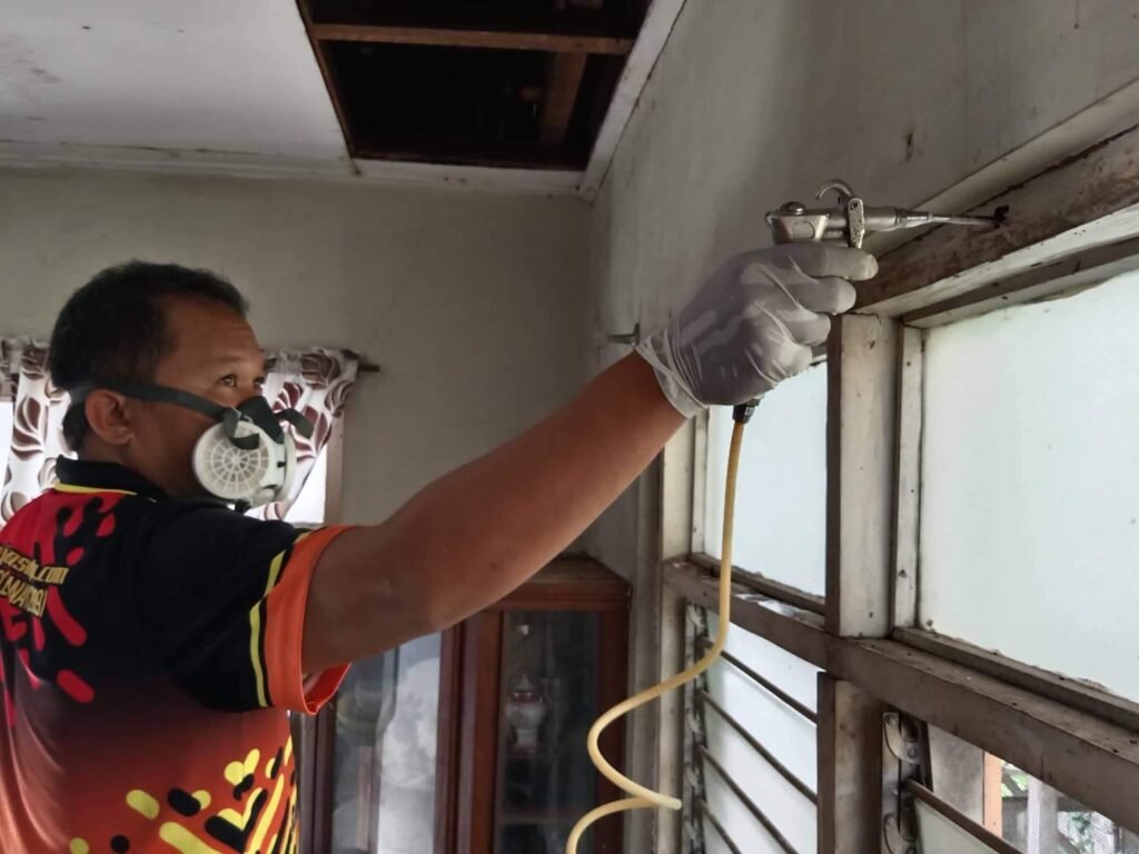 A pest control technician wearing safety gear sprays treatment into window frames inside a house.